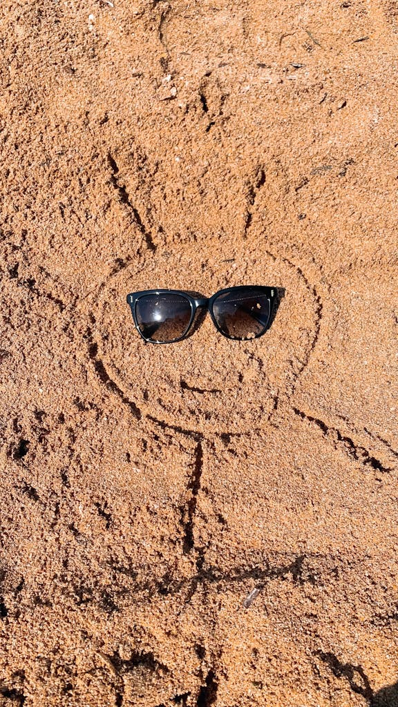 Sunglasses on a sun-shaped drawing in sandy beach texture, evoking summer vibes.