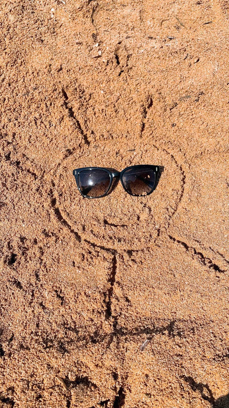 Sunglasses on a sun-shaped drawing in sandy beach texture, evoking summer vibes.
