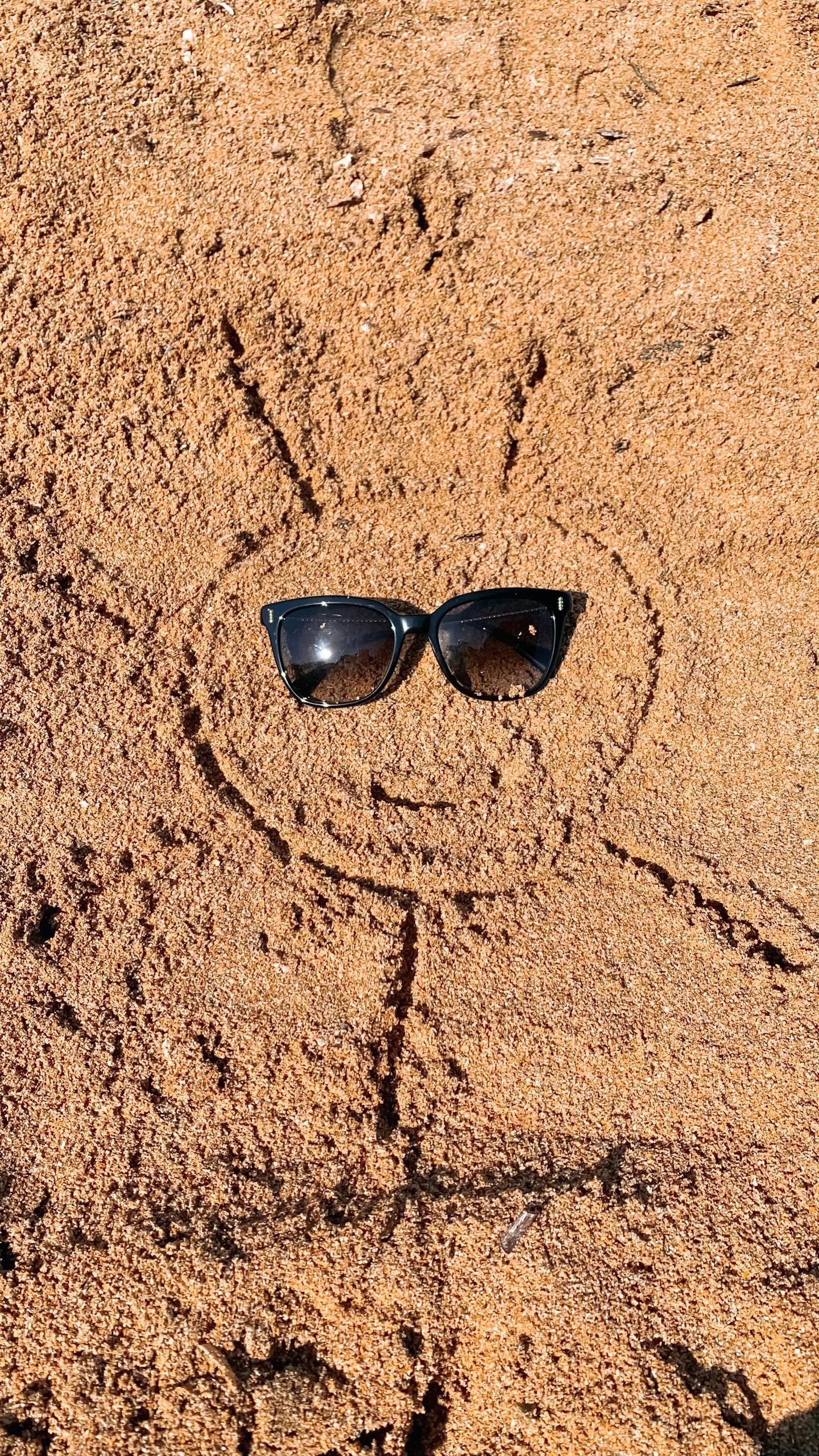 Sunglasses on a sun-shaped drawing in sandy beach texture, evoking summer vibes.